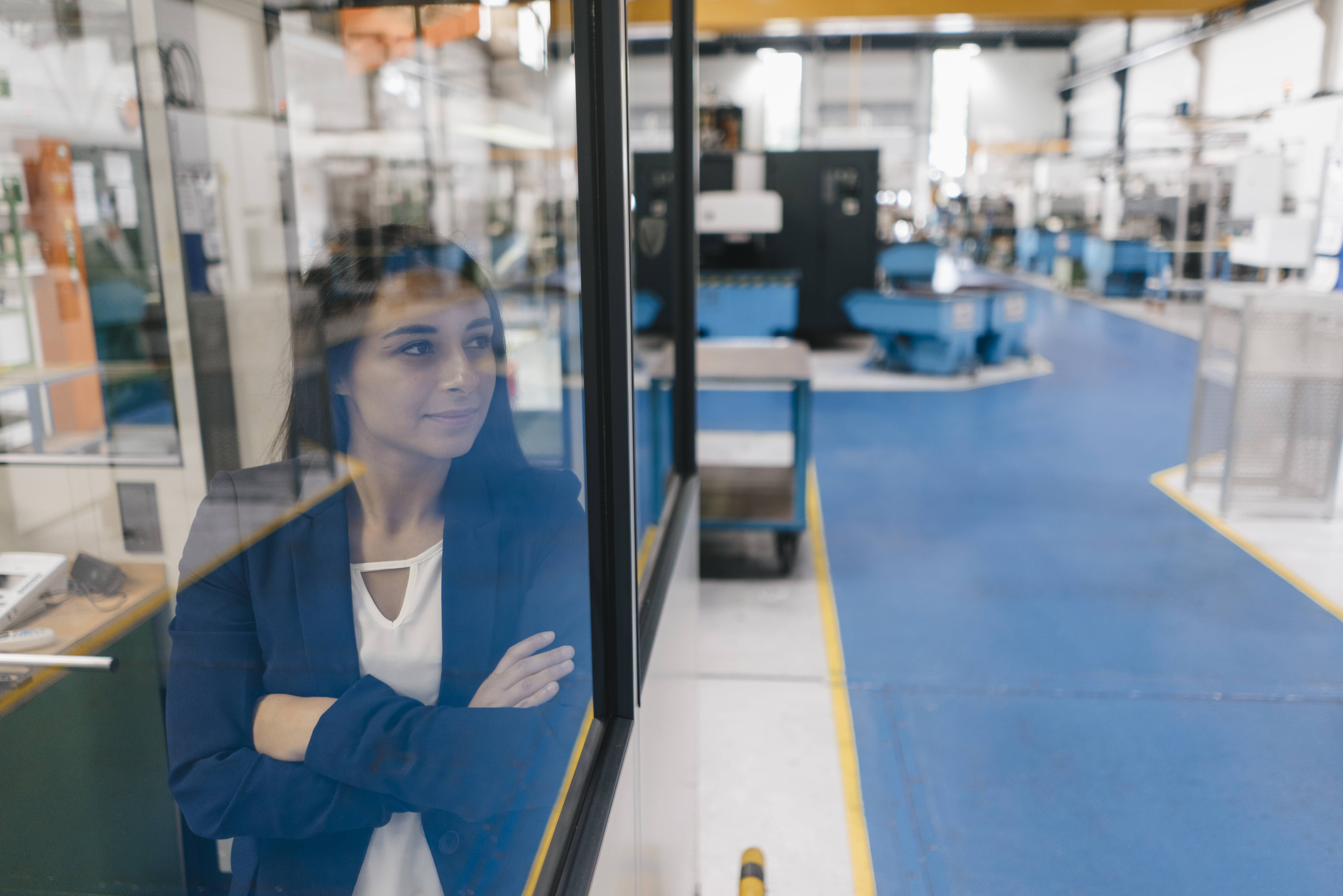 Eine Frau blickt durch eine Glasscheibe in eine High-Tech-Produktionshalle.