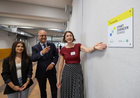 Schülersprecherin Salwa Al Ismaeel, Staatssekretär Dr. Urban Mauer und Schulleiterin Birte Hahn betrachten die neue Startchancen-Plakette an einer Wand in der Schule.