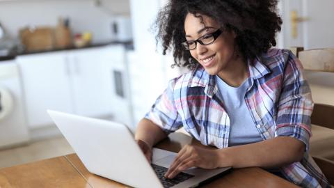 Eine Frau sitzt an einem Tisch und blickt in einen geöffneten Laptop.