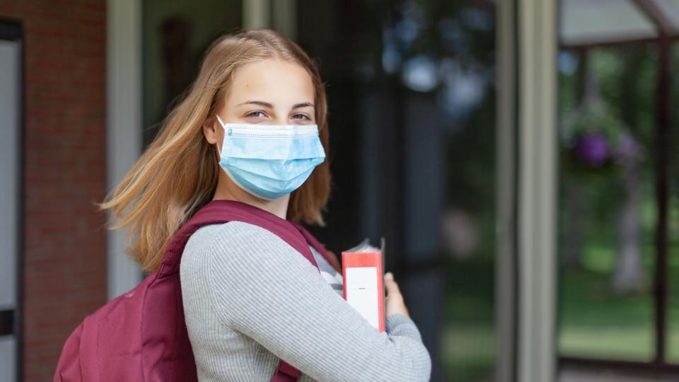 Eine Schülerin mit Maske trägt ihren Schulrucksack auf dem Rücken und einen Ordner in der Hand und blickt in die Kamera Eine Schülerin mit Maske trägt ihren Schulrucksack auf dem Rücken und einen Ordner in der Hand und blickt in die Kamera