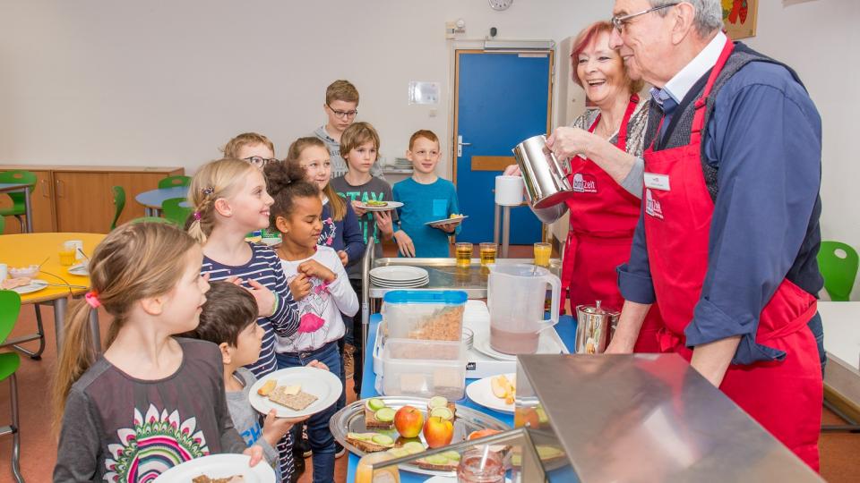 Kinder stehen vor einem Tresen, an dem zwei Erwachsene Frühstück ausgeben.