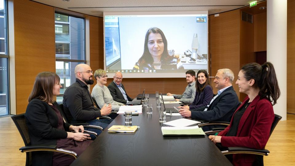 Personen sitzen in einem Besprechungsraum um einen Tisch, wobei über eine Leinwand eine externe Besprechungsteilnehmerin eingeblendet wird