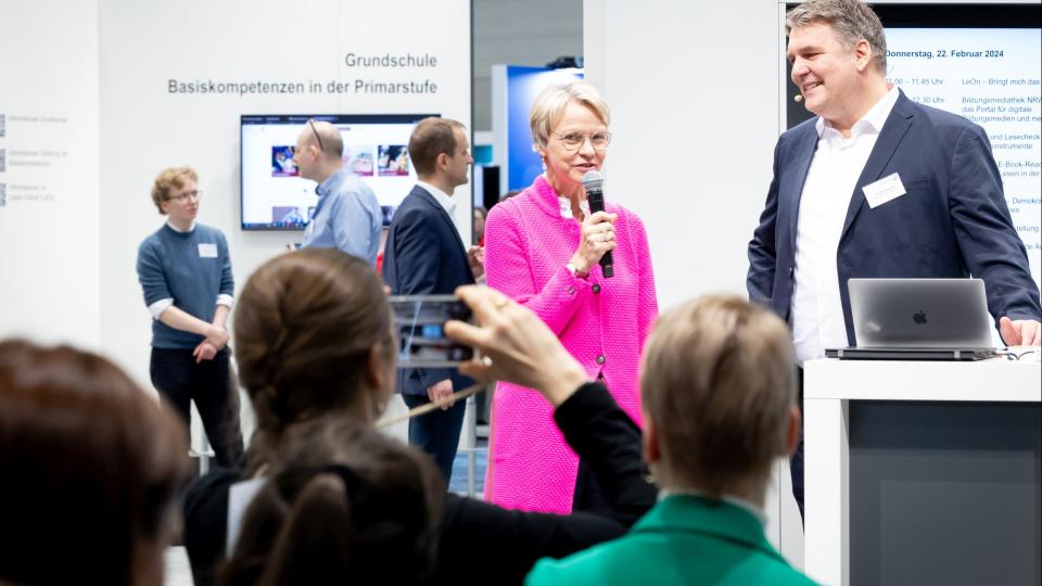 Ministerin Feller steht mit einem Mikrofon in der Hand auf der Aktivfläche des Schulministeriums bei der Vorstellung von Leon NRW, im Vordergrund Besucherinnen und Besucher der didacta 2024.