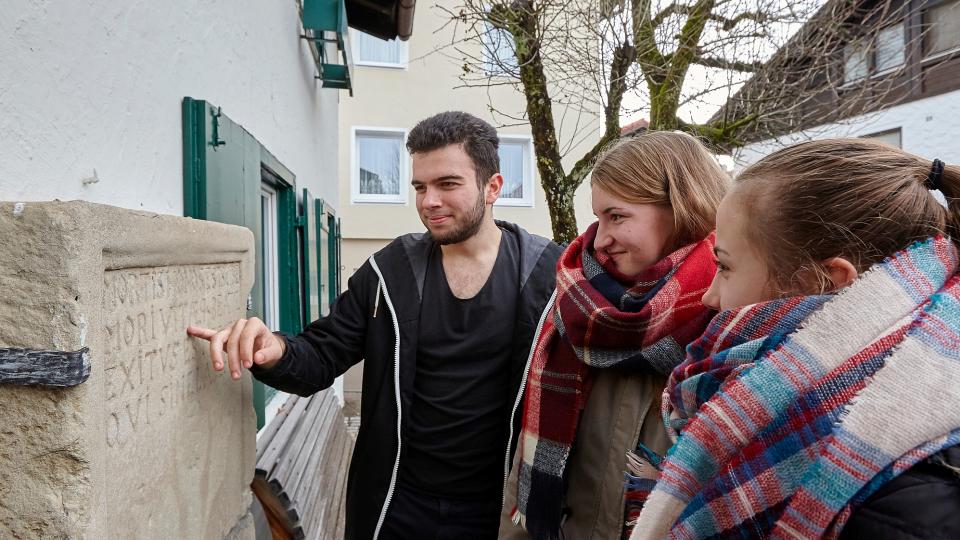 Drei  Jugendliche entziffern die Inschrift auf einem Epitaph.