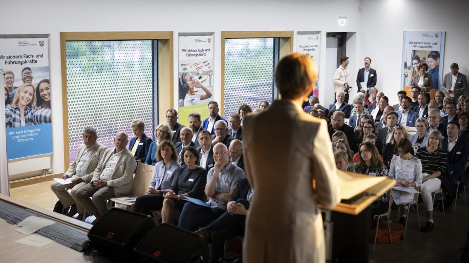 Rückansicht von Ministerin Feller, die vor Publikum bei der Festveranstaltung zum 25-jährigen Jubiläum der Berufskollegs spricht.