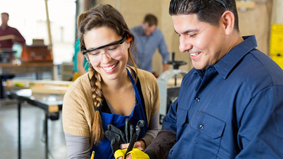 Eine Schülerin und ein Lehrer stehen zusammen in einer Werkstatt, sie tragen Arbeitskleidung und Schutzbrillen.