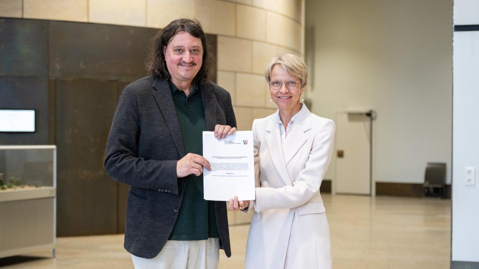 Ministerin Dorothee Feller und Prof. Dr. Christoph Selter stehen nebeneinander und halten den Zuwendungsbescheid für das Projekt PIK-AS in den Händen.