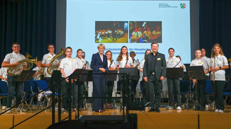 Hauptschultag in Essen: Ministerin Dorothee Feller steht mit Schülerinnen und Schülern auf einer Bühne. Die Jugendlichen halten Musikinstrumente in den Händen.