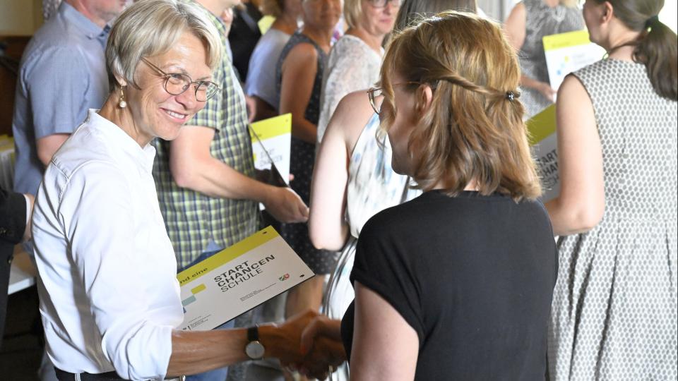 Ministerin Dorothee Feller überreicht einer Schulleiterin eine Startchancen-Plakette.