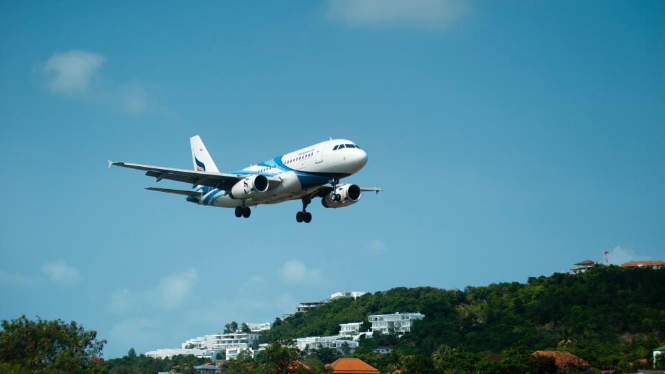 Blauer Himmel, davor startendes oder landendes Flugzeug, im Hintergrund bewaldete Landschaft mit weißen Häusern. Blauer Himmel, davor startendes oder landendes Flugzeug, im Hintergrund bewaldete Landschaft mit weißen Häusern.