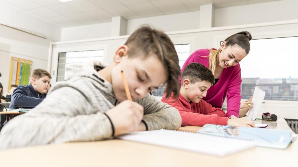 Unterrichtssituation: zwei schreibende Schüler, eine Lehrerin beugt sich über einen der beiden.