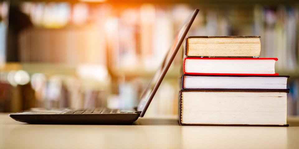 Geöffneter Laptop neben einem Bücherstapel, im Hintergrund ein Bücherregal. Geöffneter Laptop neben einem Bücherstapel, im Hintergrund ein Bücherregal.