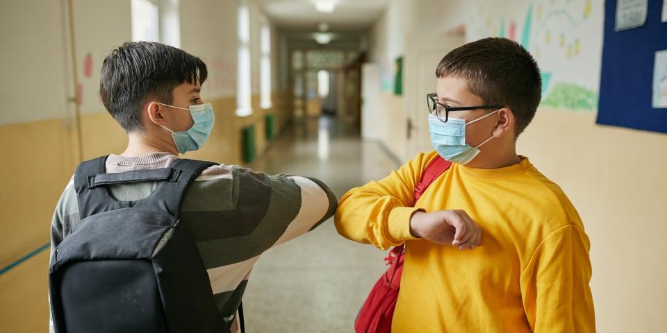 Zwei Alltagsmasken tragende Schüler begrüßen sich mit einem Ellenbogencheck auf einem Schulflur. Zwei Alltagsmasken tragende Schüler begrüßen sich mit einem Ellenbogencheck auf einem Schulflur.