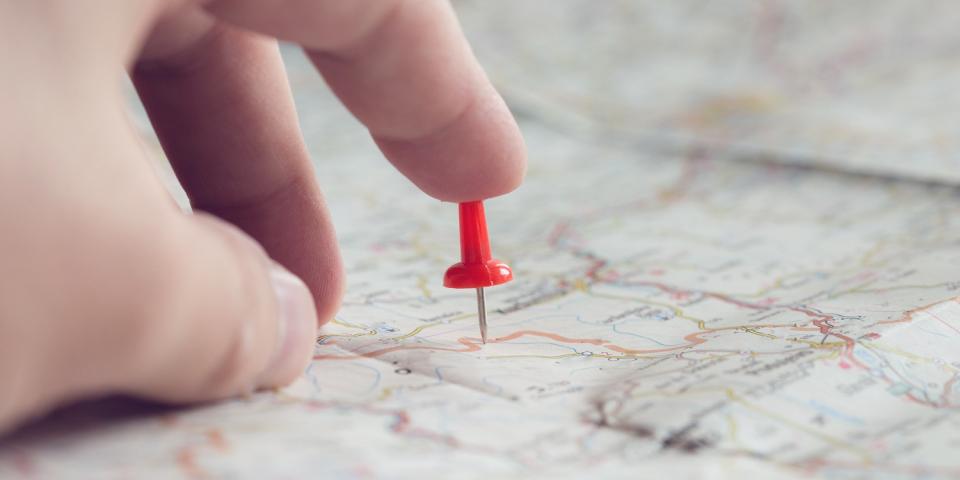 Nahaufnahme einer Landkarte, eine Hand deutet auf einen in die Karte gesteckten Pin. Nahaufnahme einer Landkarte, eine Hand deutet auf einen in die Karte gesteckten Pin.