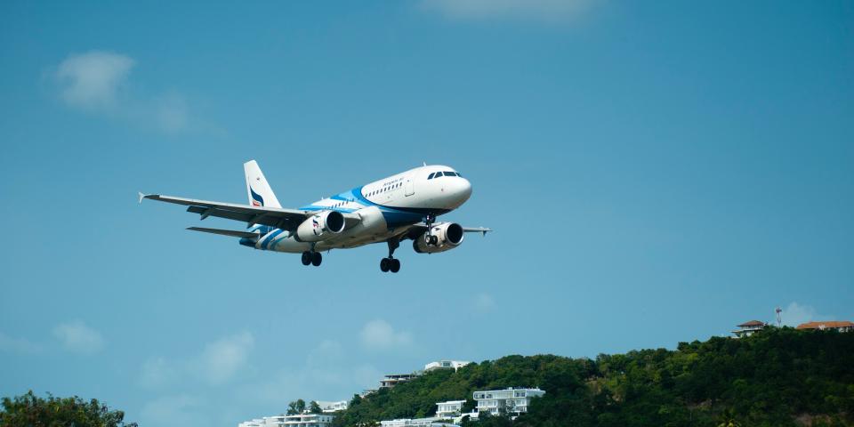 Blauer Himmel, davor startendes oder landendes Flugzeug, im Hintergrund bewaldete Landschaft mit weißen Häusern. Blauer Himmel, davor startendes oder landendes Flugzeug, im Hintergrund bewaldete Landschaft mit weißen Häusern.