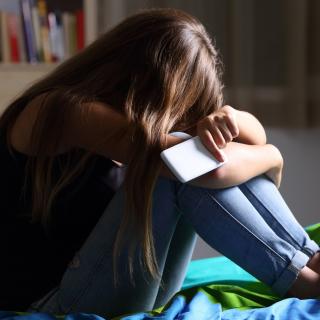 Mädchen hockt auf einem Bett, den Kopf auf die Knie gestützt, in der Hand ein Smartphone.