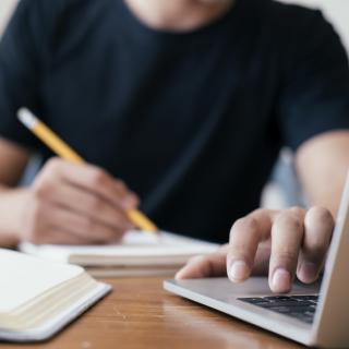 Eine Person, deren Gesicht nicht zu erkennen ist, sitzt an einem Schreibtisch, mit der einen Hand schreibend, die andere Hand liegt auf der Tastatur eines Laptops.