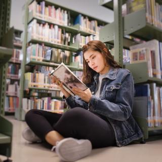 Eine Jugendliche sitzt lesend an ein Regal gelehnt in einer Bibliothek.