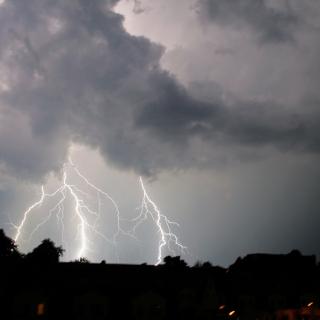 Wolkenverhangener Himmel über einer dunkel zu erkennenden Stadt. Aus den Wolken schlagen mehrere Blitze nieder.