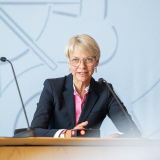 Ministerin Dorothee-Feller sitzt sprechend an einem Tisch in der Landespressekonferenz im Düsseldorfer Landtag. Im Vordergrund ist unscharf die Rückenansicht einer Person zu erkennen.