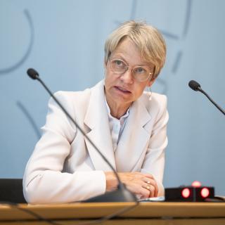 Ministerin Dorothee Feller sitzt an einem Tisch hinter zwei Mikrofonen und spricht.