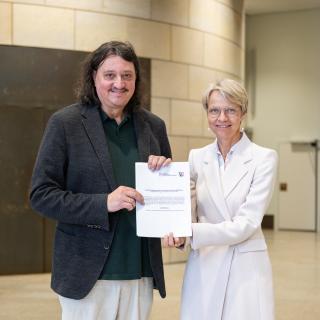 Ministerin Dorothee Feller und Prof. Dr. Christoph Selter stehen nebeneinander und halten den Zuwendungsbescheid für das Projekt PIK-AS in den Händen.