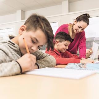 Lehrerin erklärt einem Schüler Unterrichtsstoff in einer Unterrichtssituation. Im Vordergrund sitzt ein anderer, schreibender Schüler. Lehrerin erklärt einem Schüler Unterrichtsstoff in einer Unterrichtssituation. Im Vordergrund sitzt ein anderer, schreibender Schüler.